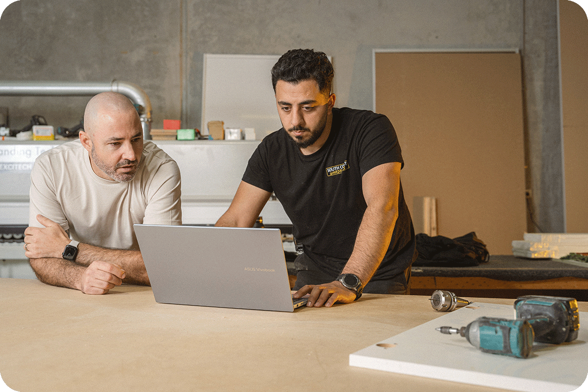 Two men are collaborating in a workshop, reviewing something on a laptop placed on a wooden table, with tools and equipment visible in the background.