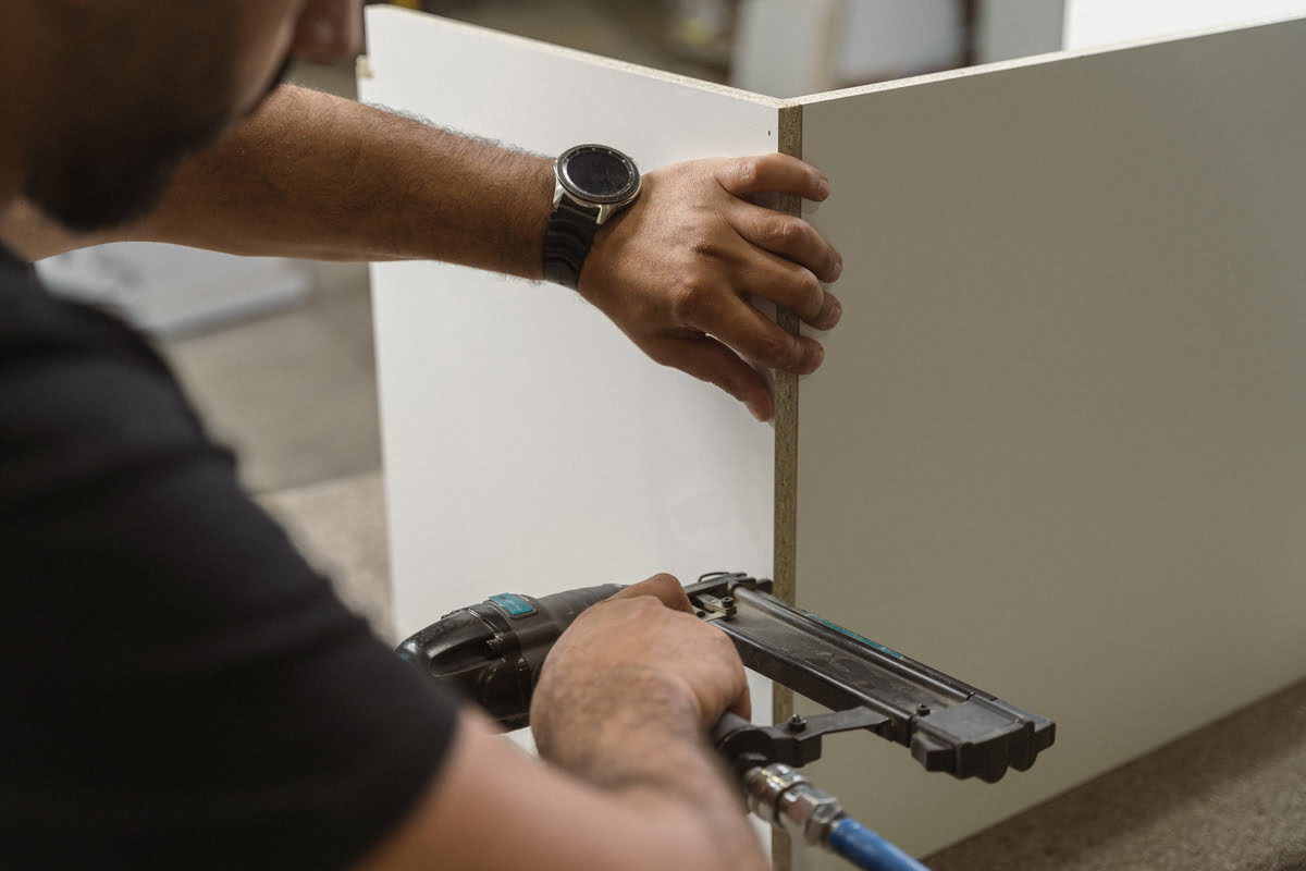 A man employs a nail gun to attach a white cabinet